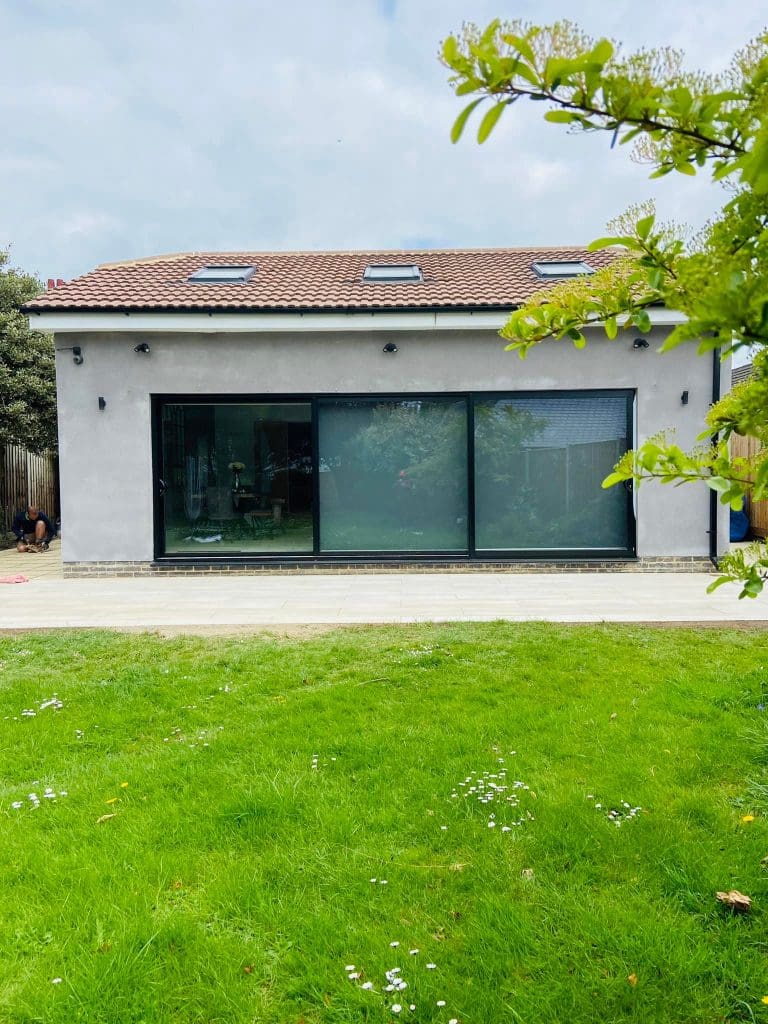 A modern house with a red-tiled roof, light grey exterior, large black-framed sliding glass doors, and skylights, seen from a green lawn with a tree branch in the foreground.