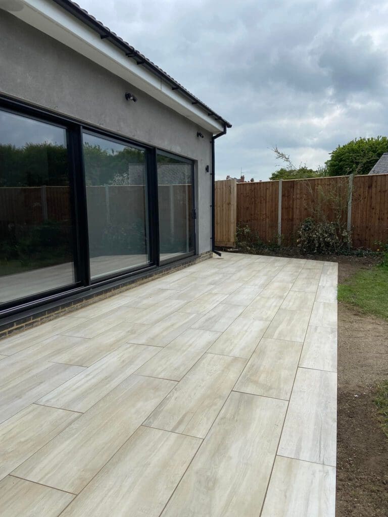 A modern patio with large, light-colored tiles next to a house with large glass doors, bordered by a wooden fence and some greenery under a cloudy sky.