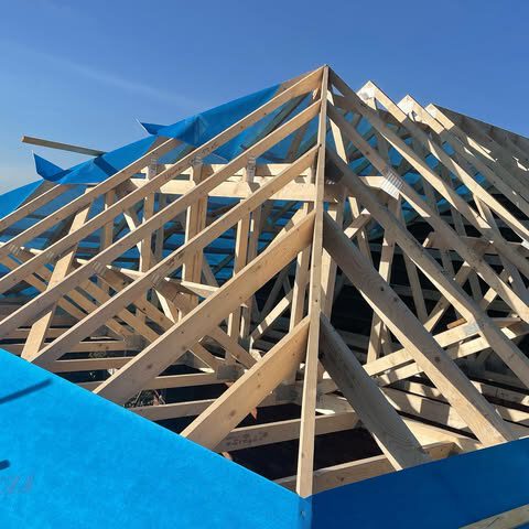 Wooden roof trusses under construction with blue tarp partially covering the structure, set against a clear blue sky.