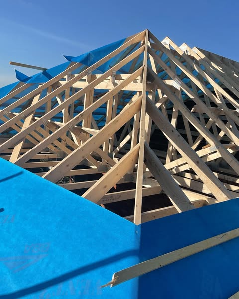A house under construction shows exposed wooden roof trusses and beams, partially covered with bright blue tarps, against a clear blue sky.