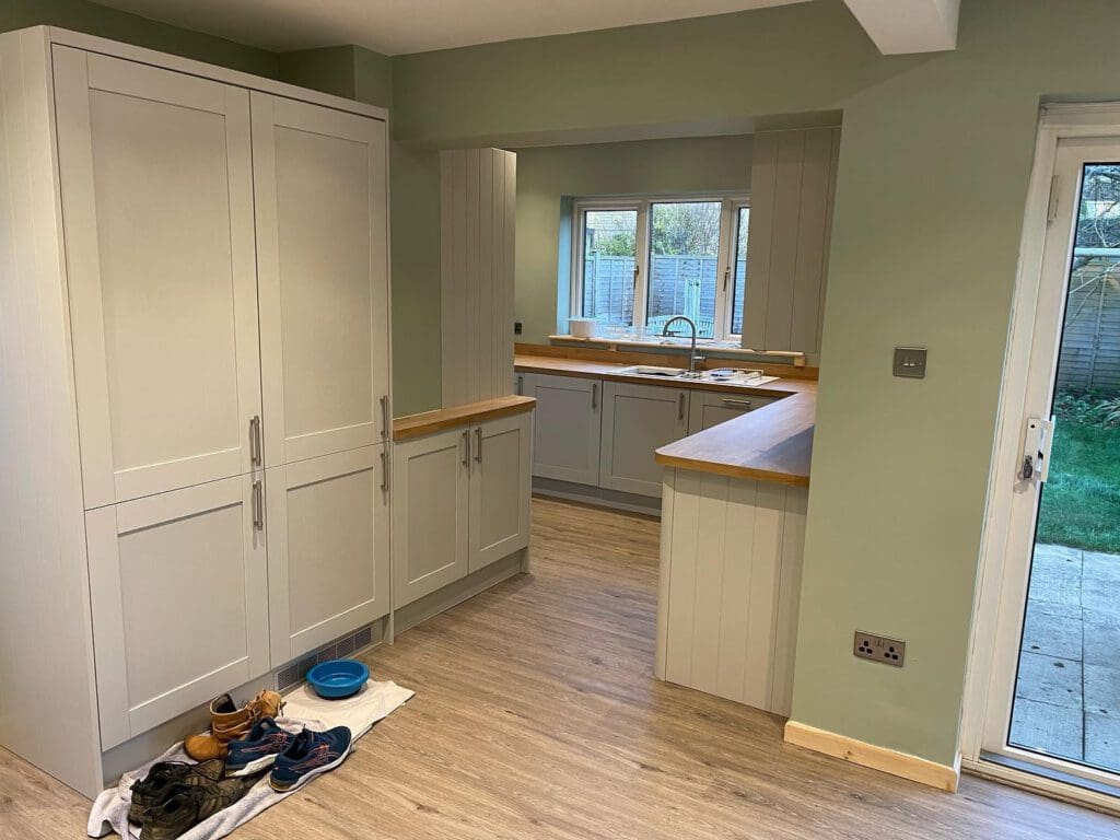 Modern kitchen with light gray cabinets, wooden countertops, and green walls. Dog bowls and shoes are on a towel on the floor. A window and glass door reveal a view of the fenced backyard.