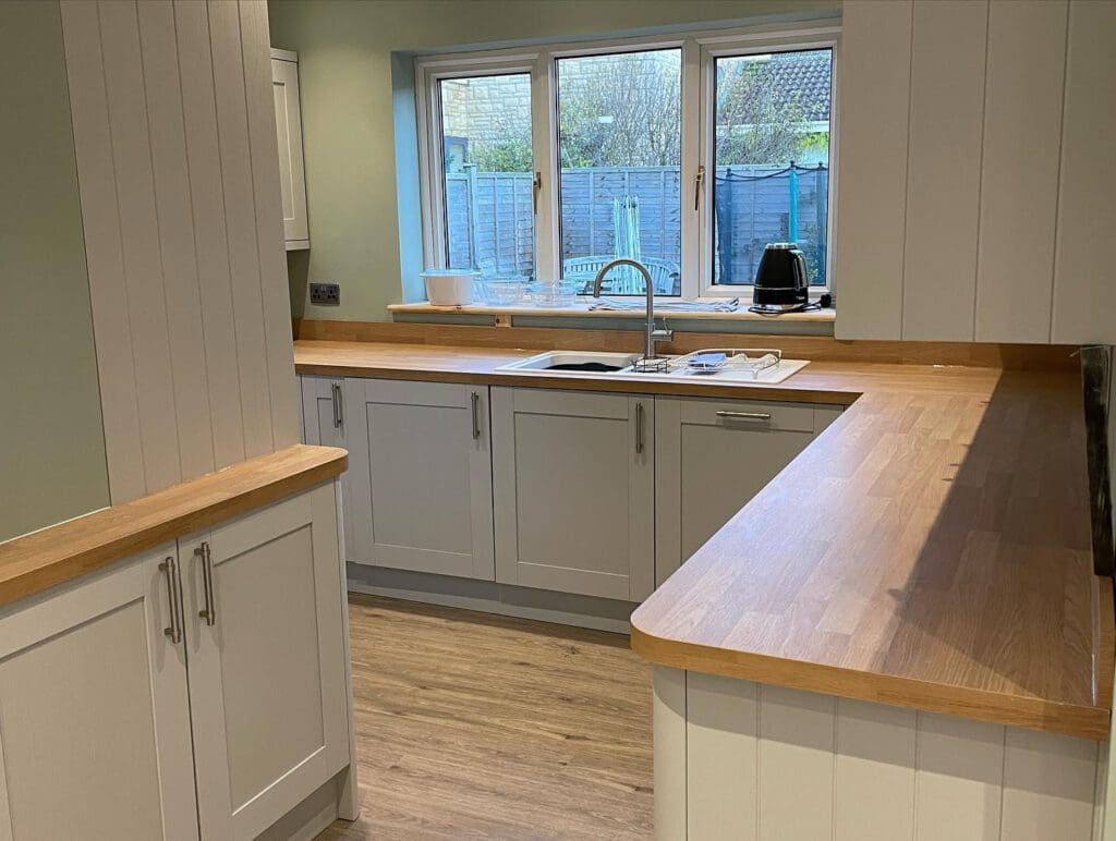 Modern kitchen with light wood countertops, white cabinets, a stainless steel sink under a window, and a few small appliances on the counters. The floor is wood, and a garden is visible through the window.