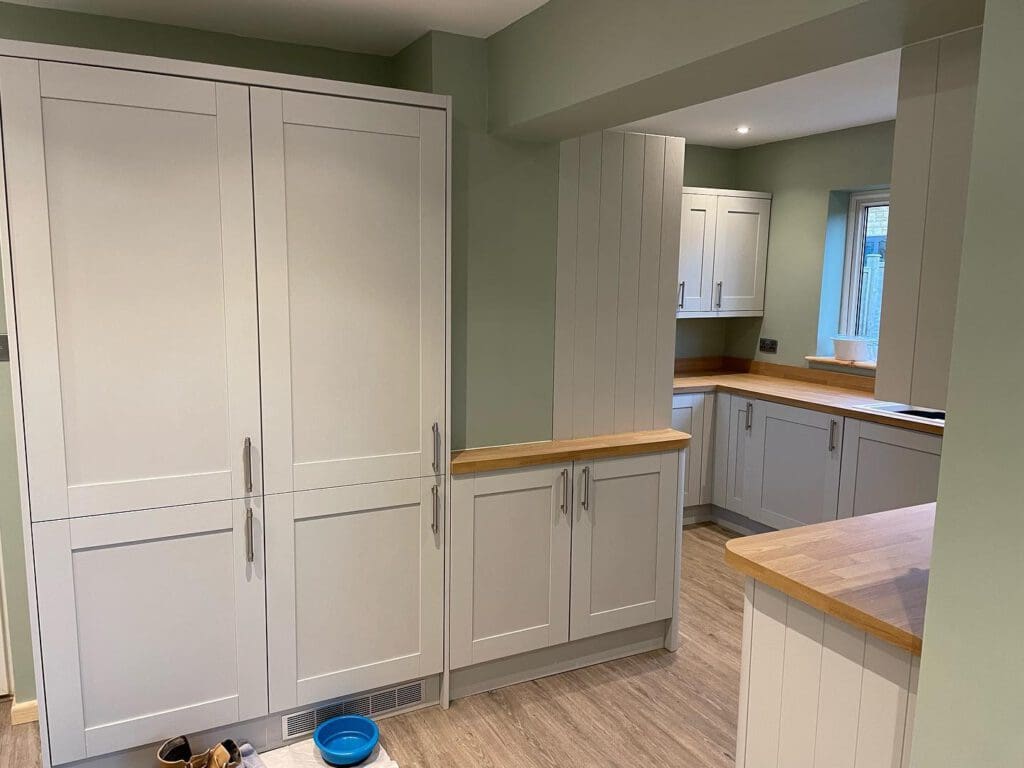 Modern kitchen with light gray cabinets, wooden countertops, and muted green walls. There is a blue pet bowl and shoes on the wood-look floor near a tall cupboard. Natural light enters through a window in the back.