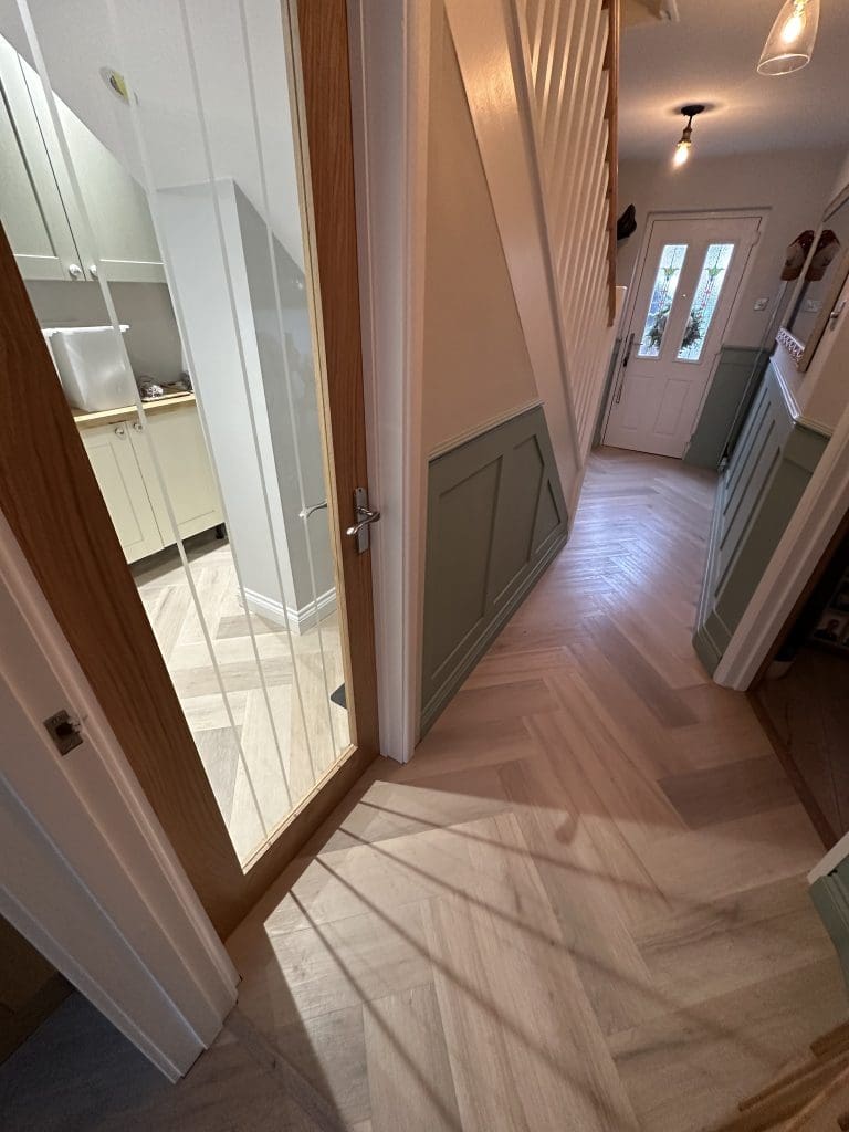 A hallway with light wood herringbone flooring, green and white paneled walls, a glass-paned door on the left leading to a utility room, and a white front door at the end with natural light coming in.
