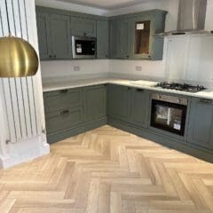 Modern kitchen with green cabinets, white countertops, built-in oven and stove, microwave, a large gold pendant light, and herringbone-patterned light wood flooring.