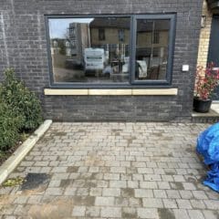 A large rectangular window set in a dark brick wall reflects parked vehicles and nearby buildings. In front of the window is a paved area bordered by plants and a blue tarp on the right.