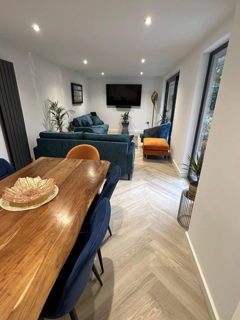 Modern living and dining room with wooden table, blue and orange chairs, teal sofas, large wall-mounted TV, plants, and light wood herringbone floor; windows line the right side, letting in natural light.