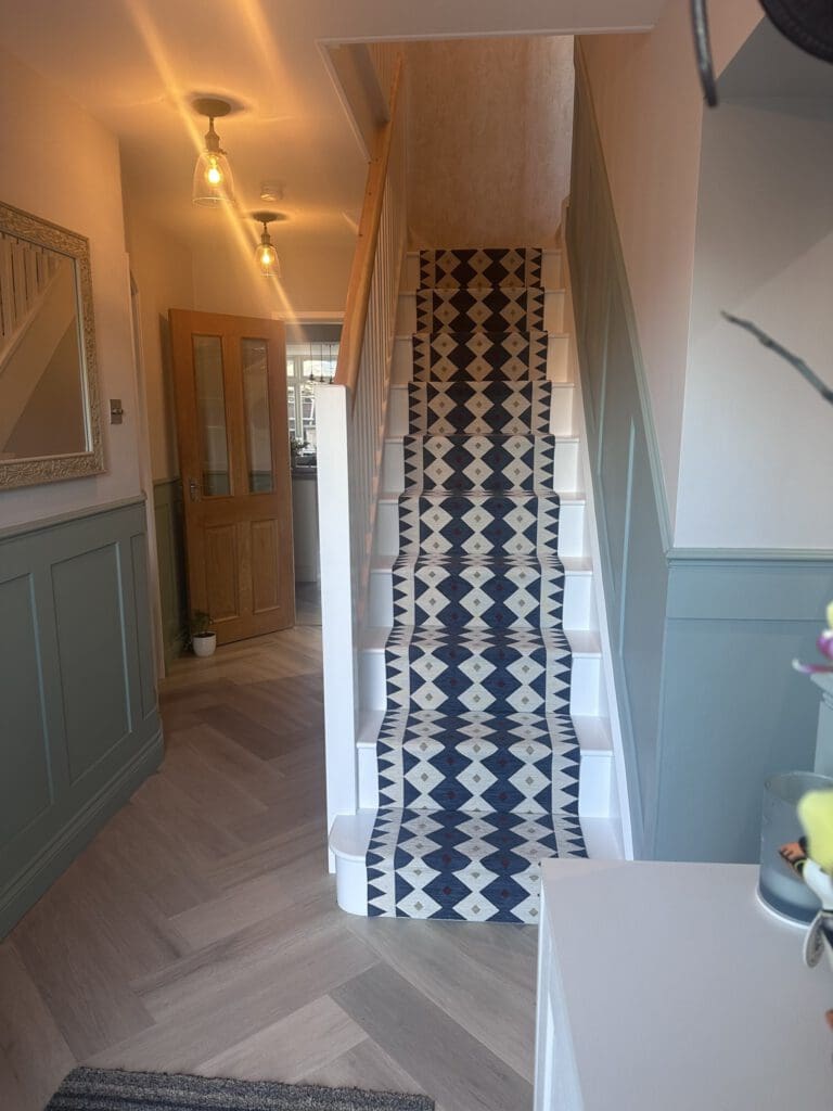 A hallway with light wood herringbone flooring, pale green wainscoting, and a staircase featuring a bold geometric-patterned runner in navy, white, and beige. A wooden door and a mirror are visible on the left.