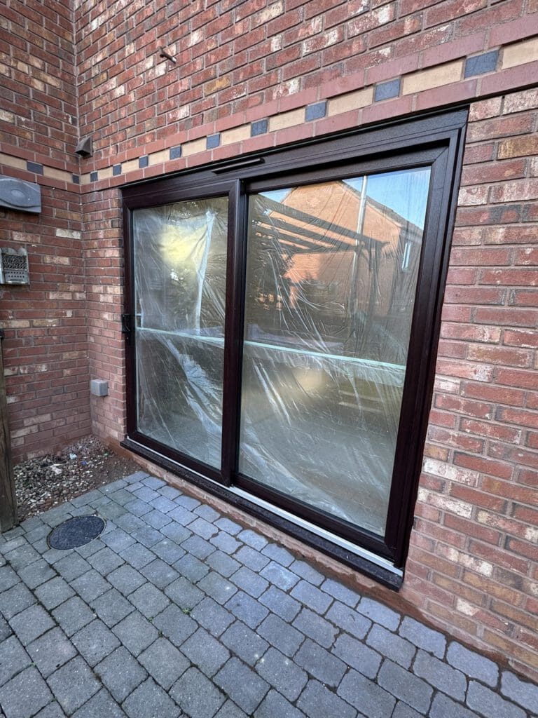 A large sliding glass door covered in protective plastic is set in a red brick wall, facing a patio with gray paving stones. The reflection of nearby houses is visible in the door.