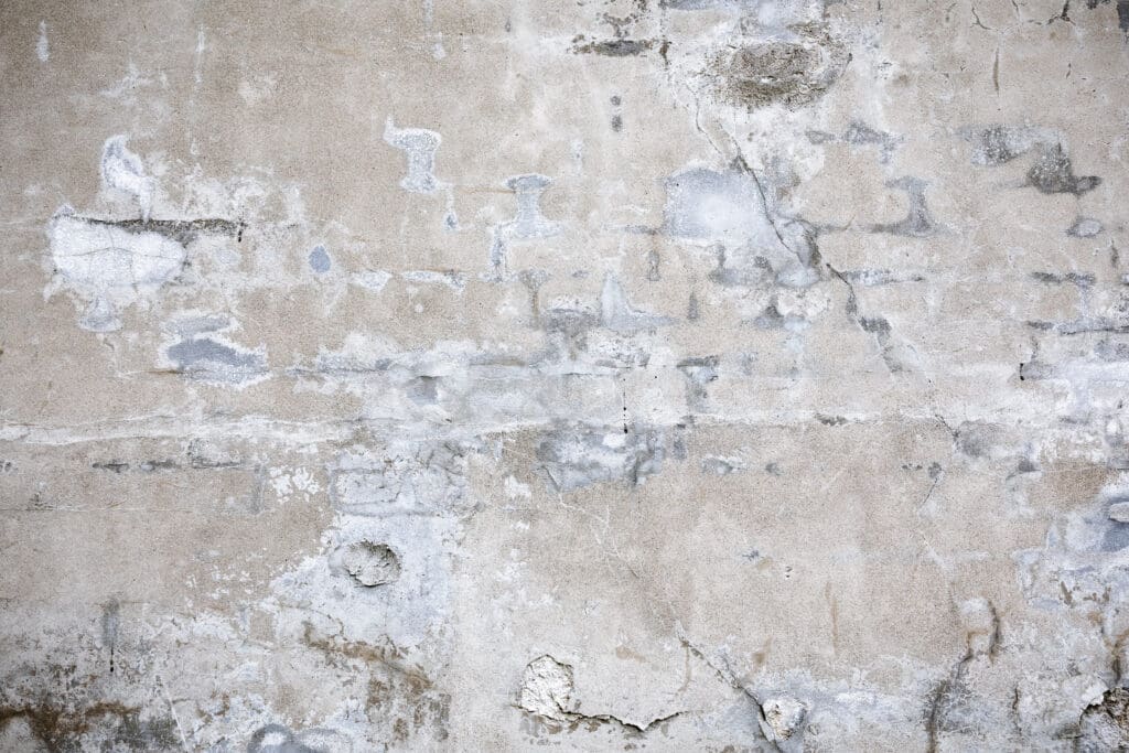 A close-up of an old, weathered concrete wall with patches, cracks, and peeling areas showing signs of aging and wear. Various shades of gray and beige create a textured, rough surface.