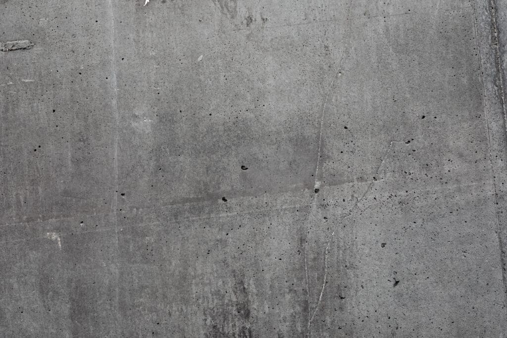 A close-up of a gray concrete wall with visible texture, small holes, cracks, and subtle stains.
