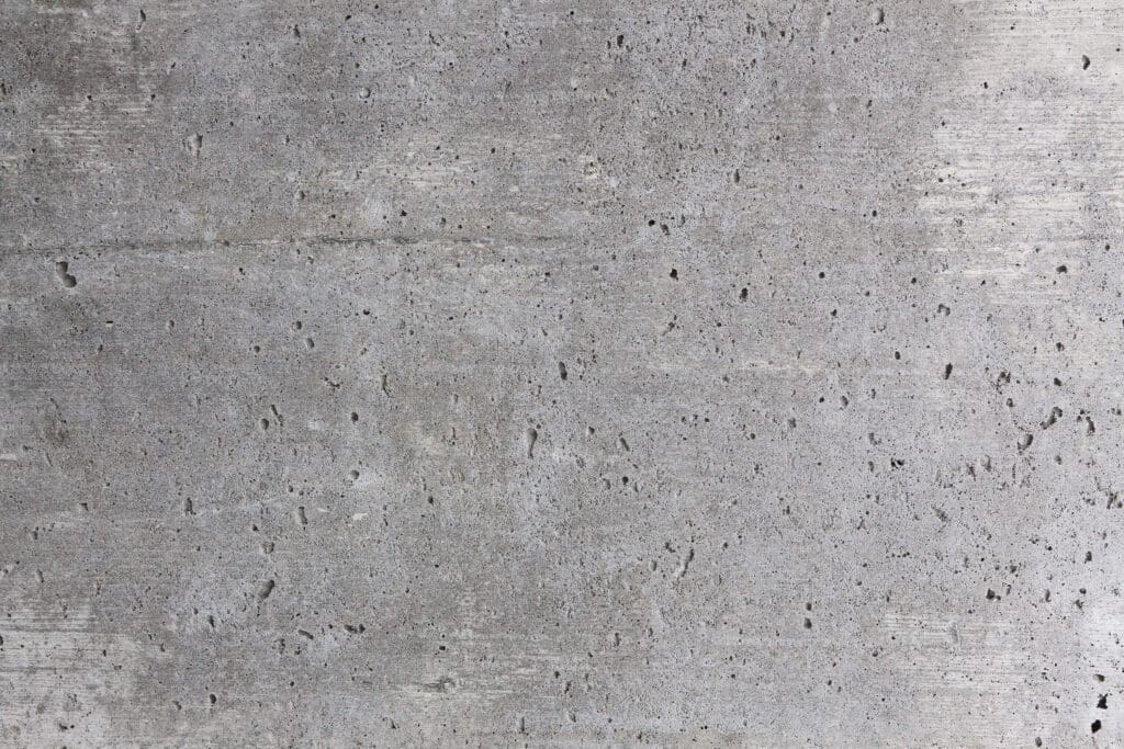 Close-up of a rough, gray concrete wall with visible small holes, pits, and a slightly uneven surface texture, giving it a raw, unfinished appearance.