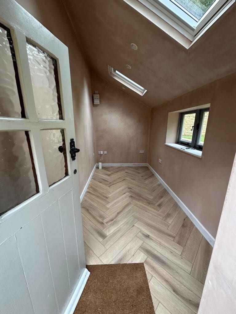 A small, empty room with herringbone wood flooring, cream-colored walls, two rectangular windows, and two skylights. The door is partially open, revealing a brown doormat just inside the entrance.