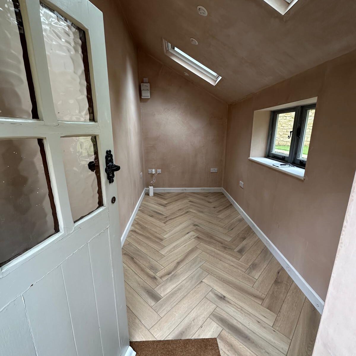 A small, narrow room with herringbone-patterned wooden flooring, light plastered walls, a white door with textured glass, a window on the right wall, and two skylights in the sloped ceiling.