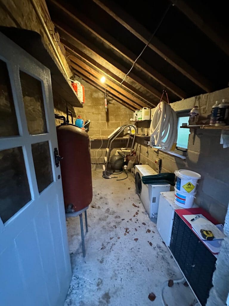 A cluttered utility room with exposed beams, a single lightbulb, a red cylinder tank, shelves with household items, and various equipment, including a water heater and washing machines, on a concrete floor.