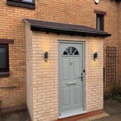 A brick porch with a light grey front door, an arched window at the top, and a mailbox slot. Two wall-mounted lights are on either side of the door, illuminating the entrance of the brick house.