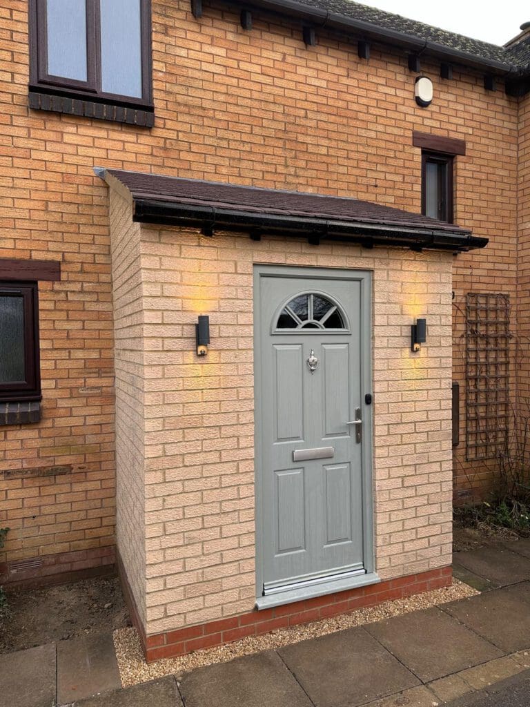 A brick porch with a light grey front door, an arched window at the top, and a mailbox slot. Two wall-mounted lights are on either side of the door, illuminating the entrance of the brick house.