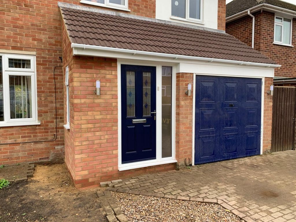 A brick house with a dark blue front door and matching garage door. The entrance has a small porch, wall lights on either side, and paved and gravel areas in front.