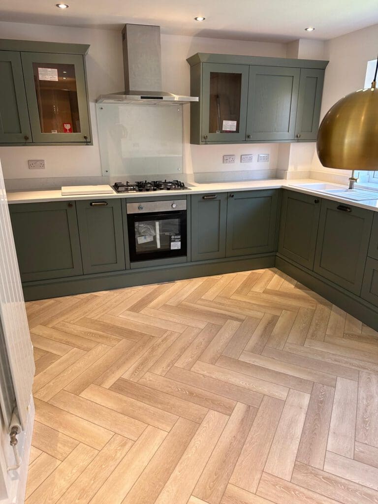 Modern kitchen with green cabinets, a built-in oven, gas stove, stainless steel hood, white countertop, and a herringbone-patterned light wood floor. A large brass pendant lamp hangs over the workspace.