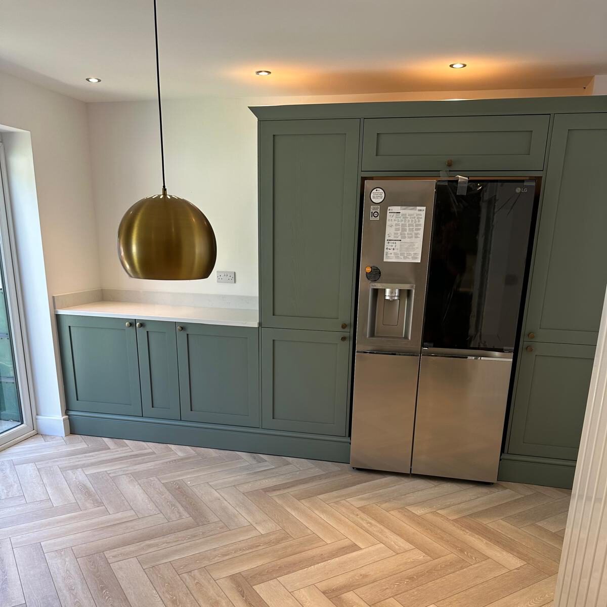 A modern kitchen with green cabinets, a stainless steel refrigerator, a brass pendant light, light wood herringbone flooring, and a counter with no appliances or clutter.