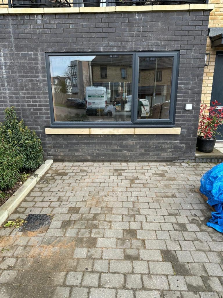 A large window set in a dark brick wall overlooks a paved area with some greenery on the left and a blue tarp on the right. Reflections of parked cars and nearby buildings are visible in the window.