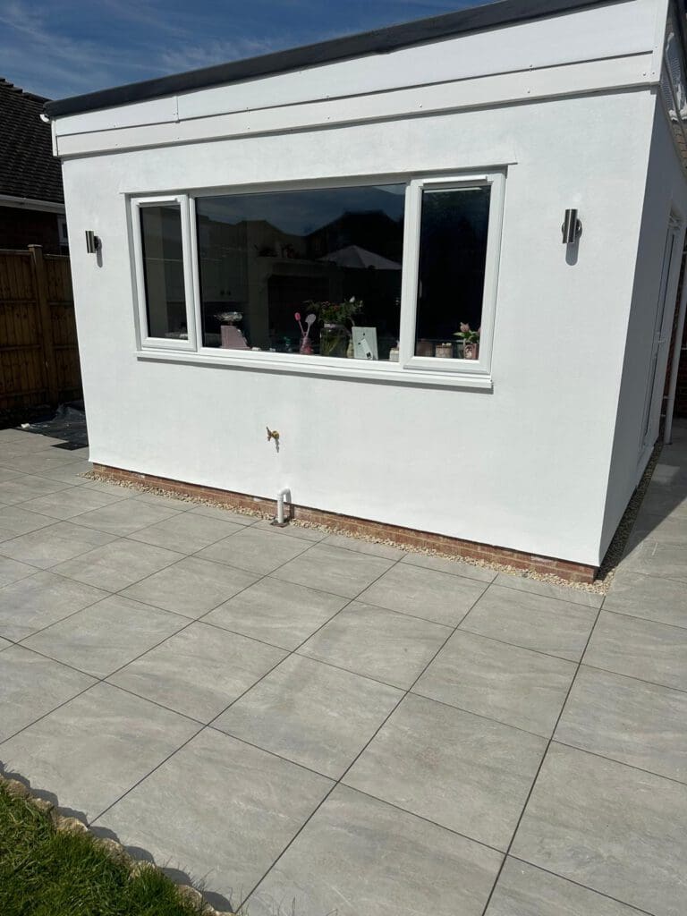 A modern white extension with a large window and outdoor wall lights, seen from a newly tiled patio under bright sunlight. Various plants and objects are visible through the window.