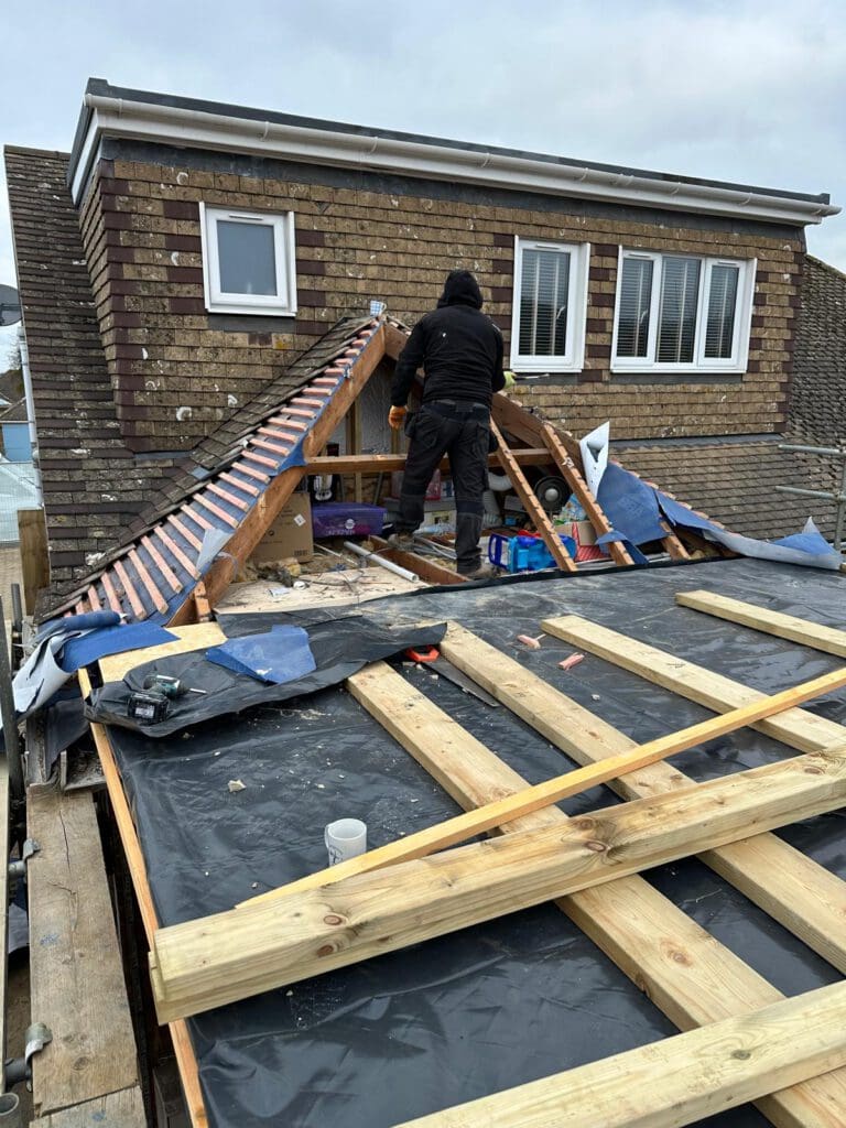 A person in dark clothing works on the wooden framework of a house roof under construction, with exposed beams, insulation, and building materials scattered around. The sky is overcast.