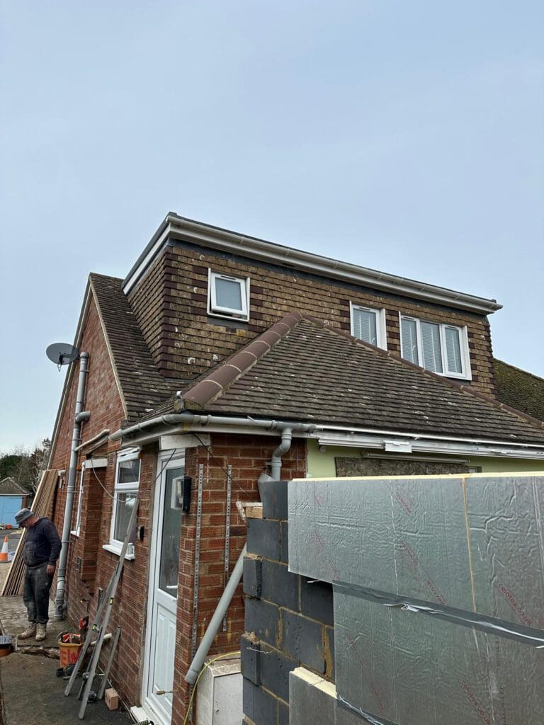 A brick house under renovation with a dormer window extension on the roof. Construction materials and insulation boards are visible in the foreground, and a person is working on the left side of the image.