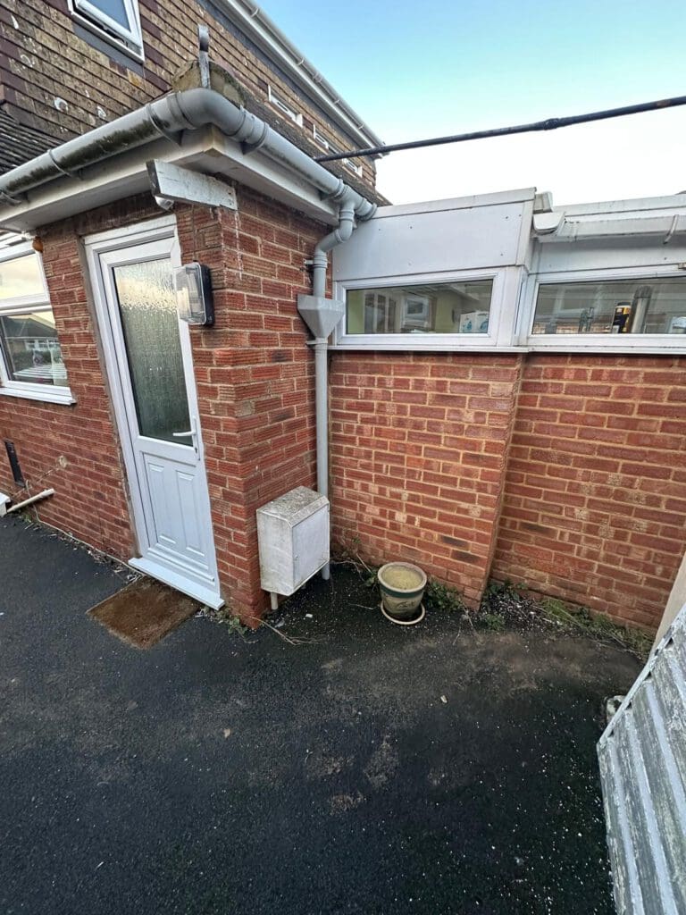 A red brick exterior wall with a white door, a drainpipe, a small utility box, a brown doormat, and a round beige plant pot on a black paved surface. There are windows above the wall and guttering along the roof.