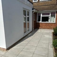A paved patio with large grey tiles next to a white extension on a brick house. A garden bed with plants lines the right edge, and there are windows on both the extension and the main house.