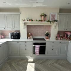 Bright kitchen with pale grey cabinets, pink subway tile backsplash, and white countertops. Decorative shelves hold plants and kitchenware. A pink stand mixer and air fryer sit on the counters. Sunlight streams in from above.
