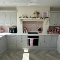 Bright kitchen with pale grey cabinets, pink backsplash tiles, and light wood herringbone floor. Pink accents, plants, and baking appliances are displayed on the counters and shelves, with sunlight streaming in from above.