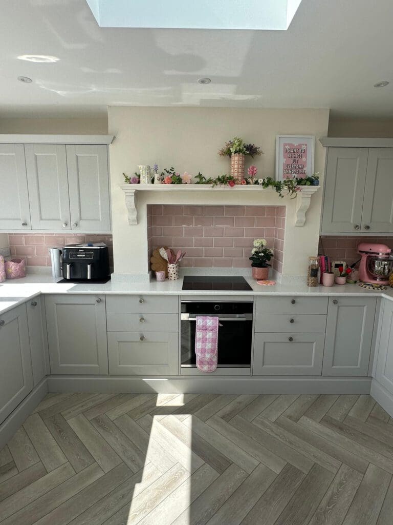 Bright kitchen with pale grey cabinets, pink backsplash tiles, and light wood herringbone floor. Pink accents, plants, and baking appliances are displayed on the counters and shelves, with sunlight streaming in from above.