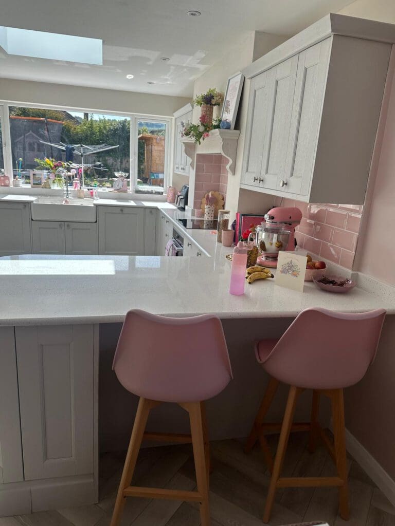 A bright kitchen with white cabinets, pink accents, and two pink barstools at a white countertop. Sunlight streams in through large windows, and various kitchen items and a card are on the counter.