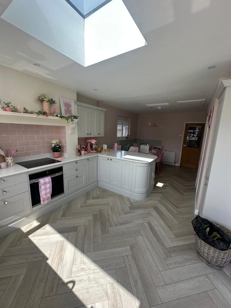 Bright, modern kitchen with pale grey cabinets, pink accents, and herringbone wood flooring. Skylight lets in natural light. Adjacent dining area with pink chairs and decor. Wicker basket on the right.