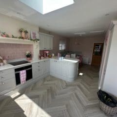 Modern kitchen with light grey cabinets, pink accents, and herringbone wood flooring. A skylight brightens the space, and a basket sits in the corner. The kitchen opens into a dining area with a table and pink chairs.