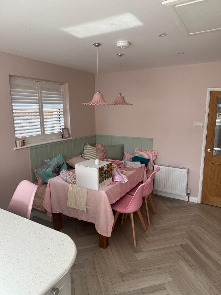 A cozy dining nook with pink walls, a corner bench decorated with pastel cushions, a pink tablecloth, pink chairs, and two pink pendant lights. A gift box and tissue paper sit on the table. Sunlight comes through window blinds.