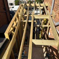 A view from above of a building extension under construction between two brick houses, showing exposed wooden beams and framing, with tools and construction materials scattered below.