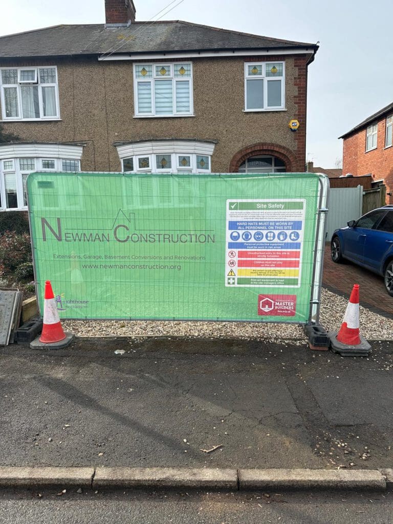 A green construction barrier with "Newman Construction" branding and site safety information stands in front of a residential house. Traffic cones are placed at each end of the barrier. A blue car is parked nearby.