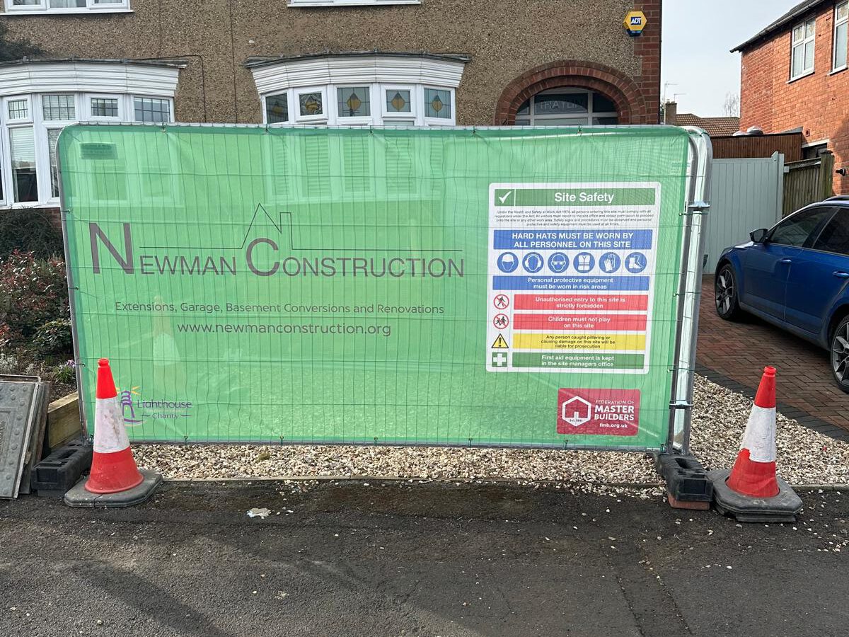 A green construction site barrier with "Newman Construction" logo and details, safety instructions, and warning signs. Two orange traffic cones are placed on the street in front of the barrier. Houses are visible in the background.