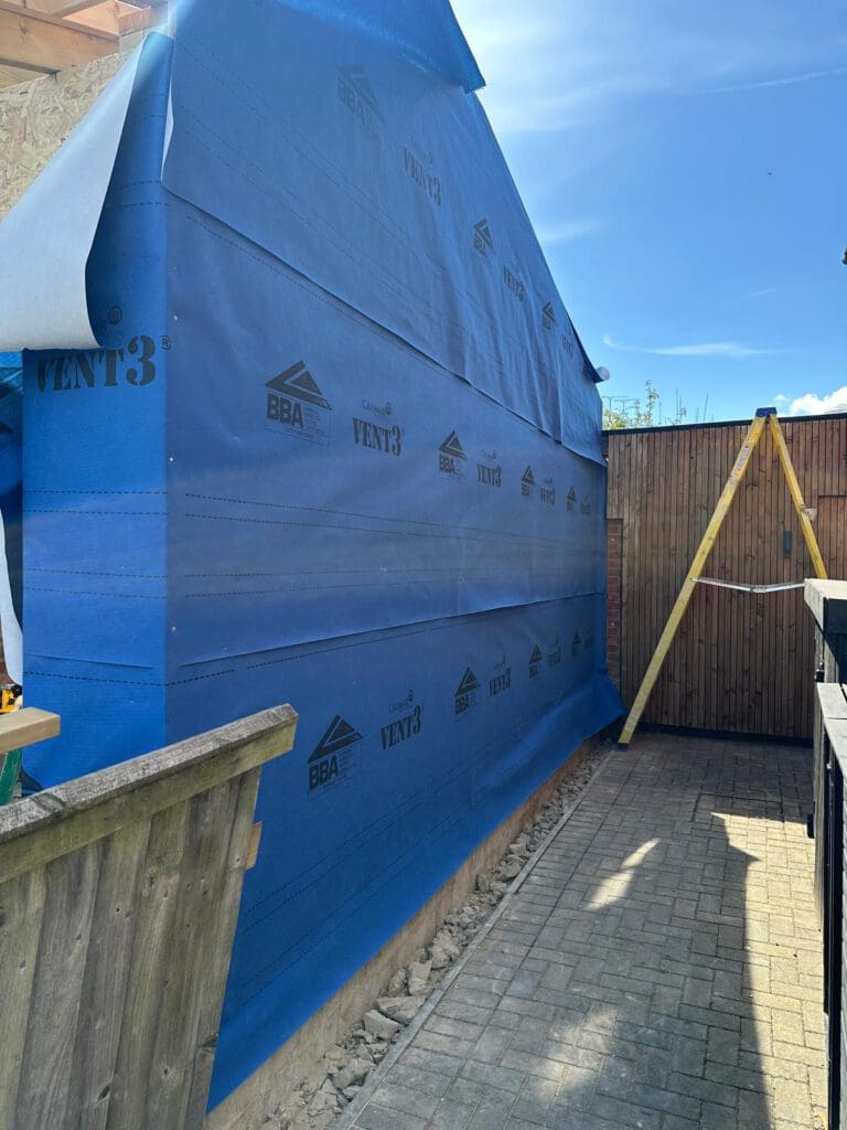 A building under construction is covered with blue weatherproof membrane labeled "VENT3" and "BBA." The area beside the building is paved with bricks, and a wooden fence and a yellow ladder are visible.