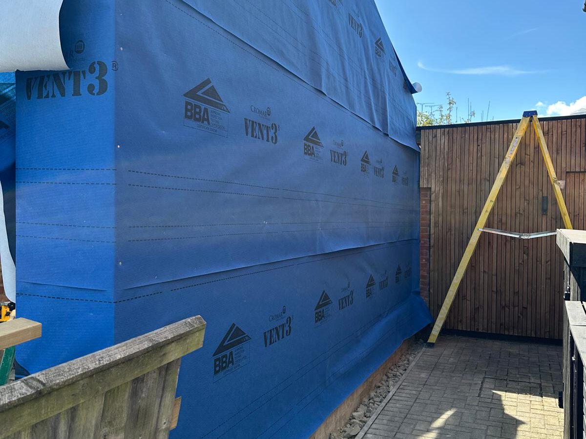 A building under construction covered with blue breathable membrane sheeting labeled "VENT3." A wooden fence and a wooden ladder are nearby, and the sky is clear and sunny.