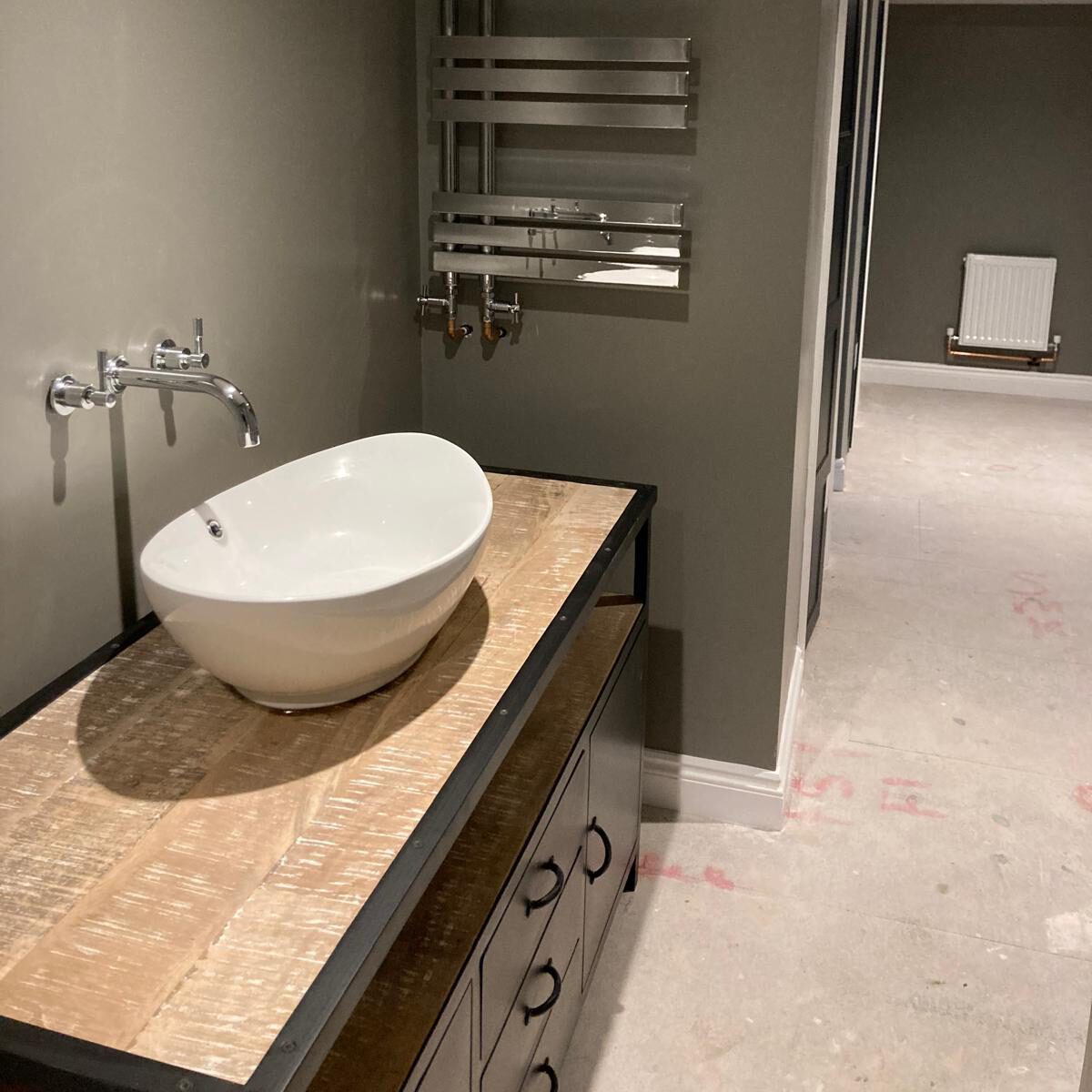 Modern bathroom with a white vessel sink on a wooden countertop, wall-mounted faucet, towel rail above, and light gray walls. The floor is unfinished, showing markings, and an open doorway leads to another room.