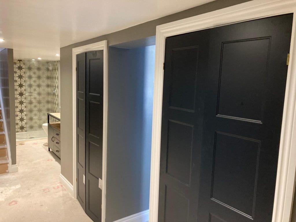 A hallway with gray walls, black paneled doors with white trim, and an unfinished floor. In the background, there is patterned wallpaper and a partially visible ladder.