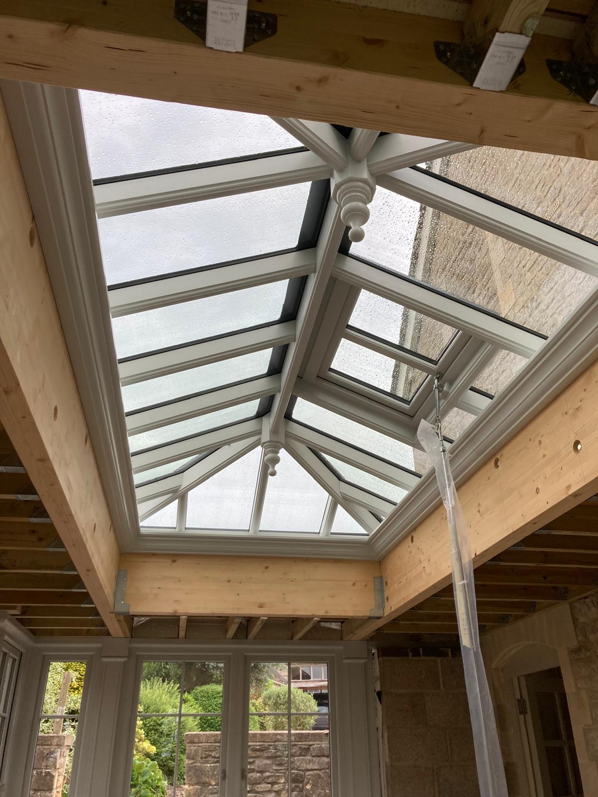 A glass roof lantern with white framing is installed in a partially constructed room with exposed wooden beams, providing a clear view of the sky and outdoor greenery.