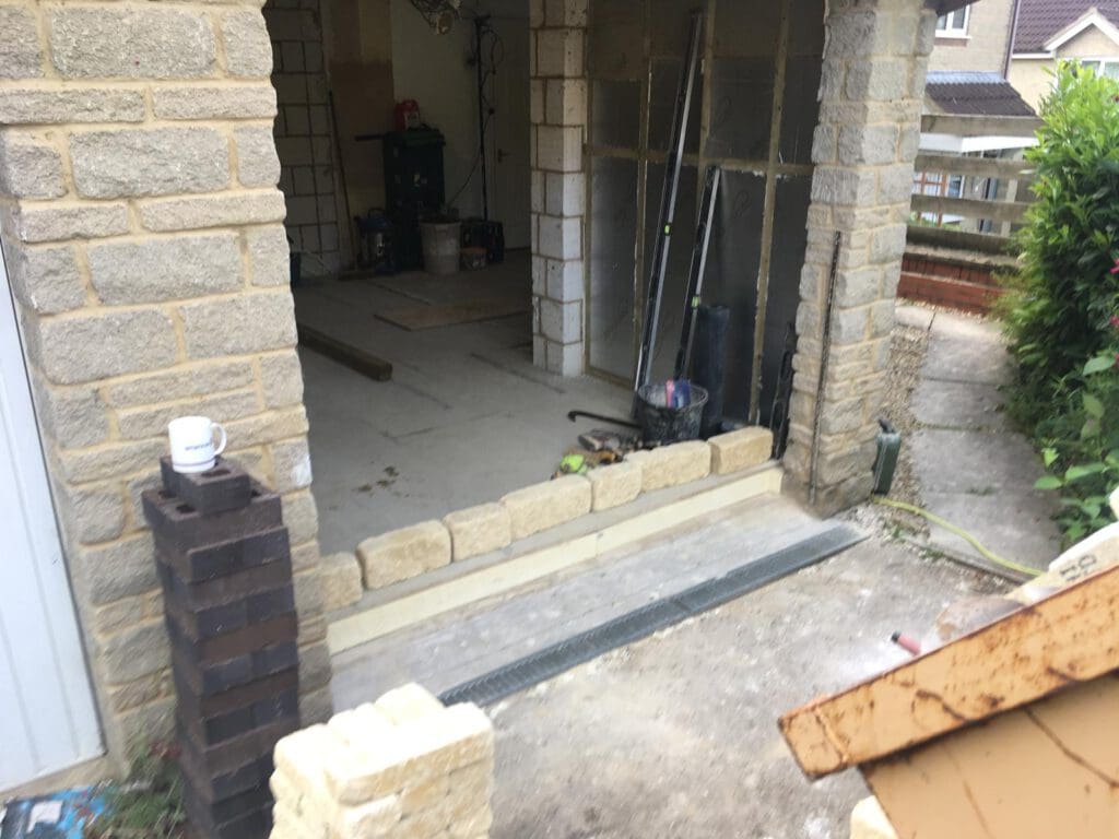 A partially built stone wall at the entrance of a house extension, with tools and building materials scattered inside and outside. A stack of bricks, a mug, and construction equipment are visible.