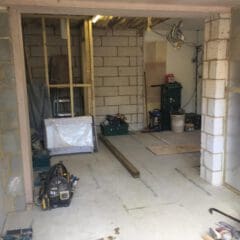 A partially constructed room with exposed brick walls, construction tools, stacked toolboxes, and building materials scattered on the floor. Wooden frames and insulation are visible, indicating ongoing renovation work.