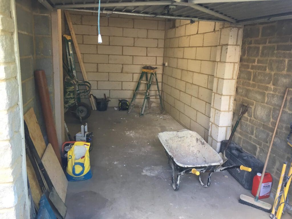 A small unfinished garage or storage room with bare brick walls, a metal roof, a wheelbarrow, folding ladder, construction tools, wooden planks, rubbish bags, and a red fuel container scattered on the concrete floor.