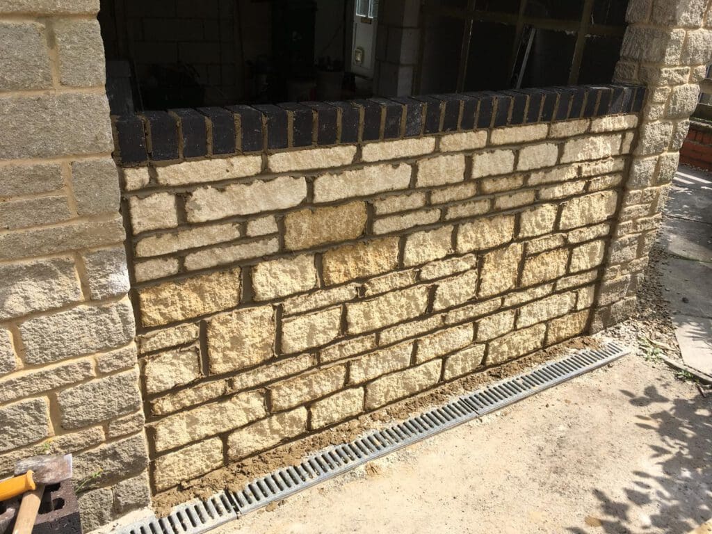 A stone wall with light-colored bricks and a row of dark bricks along the top sits above a metal drainage grate on a concrete surface, with sunlight casting shadows on the area.