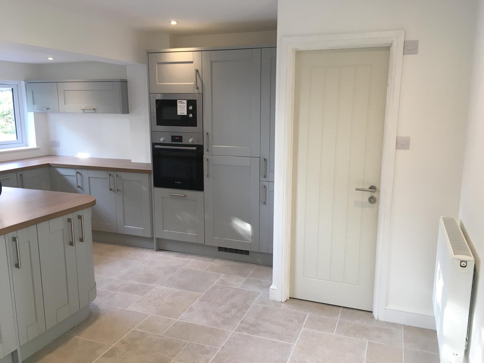 Modern kitchen with light gray cabinets, built-in oven and microwave, wooden countertops, beige floor tiles, and a white door. Wall radiator is visible on the right side. Room is bright with natural light.